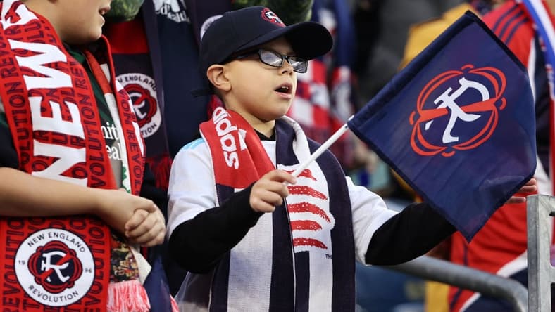 Nov 7, 2021; Foxborough, Massachusetts, USA; A young New England Revolution fan waves a flag with the team s new logo for the 2022-23 season during the first half of the game between the New England Revolution and the Inter Miami at Gillette Stadium. Mandatory Credit: Winslow Townson-USA TODAY Sports