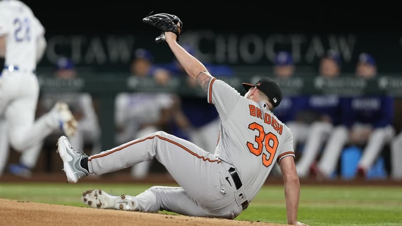 Apr 3, 2023; Arlington, Texas, USA; Baltimore Orioles starting pitcher Kyle Bradish (39) falls to the ground after getting hit by a ball off the bat of Texas Rangers catcher Jonah Heim (28) during the second inning at Globe Life Field. Mandatory Credit: Jim Cowsert-USA TODAY Sports