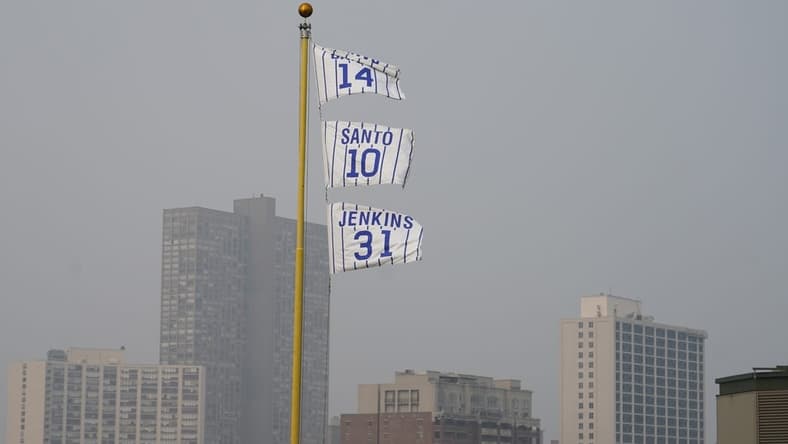Jun 27, 2023; Chicago, Illinois, USA; The flags of Chicago Cubs' Hall of Fame players Ernie Banks (14), Ron Santo (10) and Ferguson Jenkins fly off the Wrigley Field left field foul pole as a veil of haze from Canadian wildfires shroud high rise buildings along Lake Michigan before a baseball game between the Cubs and the Philadelphia Phillies at Wrigley Field. Mandatory Credit: David Banks-USA TODAY Sports
