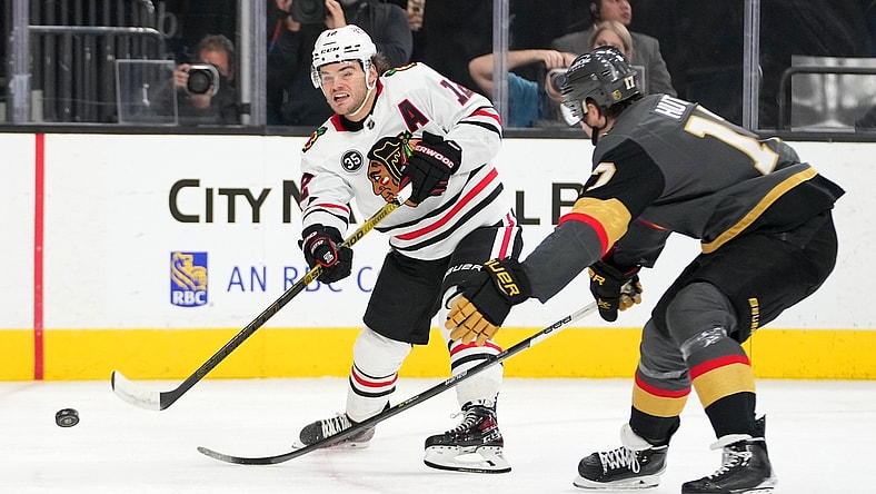 Mar 26, 2022; Las Vegas, Nevada, USA; Chicago Blackhawks left wing Alex DeBrincat (12) shoots in front of Vegas Golden Knights defenseman Ben Hutton (17) during the first period at T-Mobile Arena. Mandatory Credit: Stephen R. Sylvanie-USA TODAY Sports