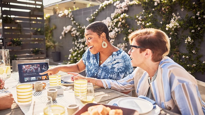 Two Girlfriends Watching Sling TV On Their Tablet While Eating On Their Patio
